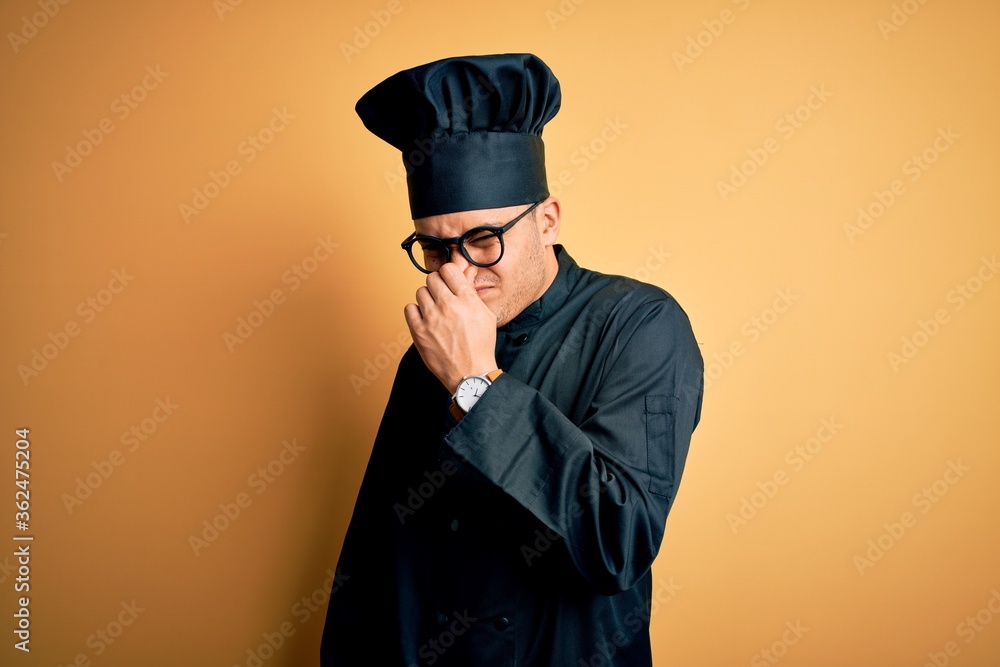 Young brazilian chef man wearing cooker uniform and hat over isolated yellow background smelling something stinky and disgusting, intolerable smell, holding breath with fingers on nose. Bad smell Young brazilian chef man wearing cooker uniform and hat over isolated yellow background smelling something stinky and disgusting, intolerable smell, holding breath with fingers on nose. Bad smell
