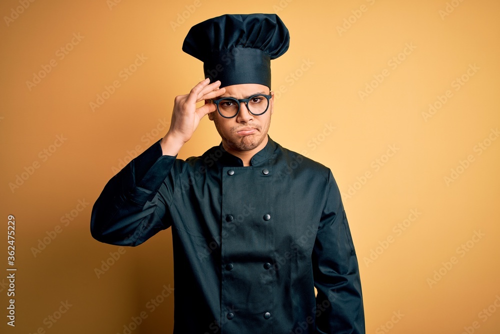 Young brazilian chef man wearing cooker uniform and hat over isolated yellow background worried and stressed about a problem with hand on forehead, nervous and anxious for crisis Young brazilian chef man wearing cooker uniform and hat over isolated yellow background worried and stressed about a problem with hand on forehead, nervous and anxious for crisis
