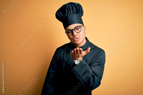 Young brazilian chef man wearing cooker uniform and hat over isolated yellow background looking at the camera blowing a kiss with hand on air being lovely and sexy. Love expression.