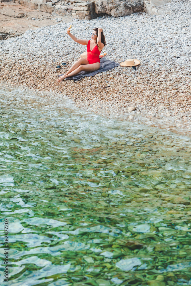 young pretty woman in red swimsuit sitting at rocky beach with phone taking selfie picture