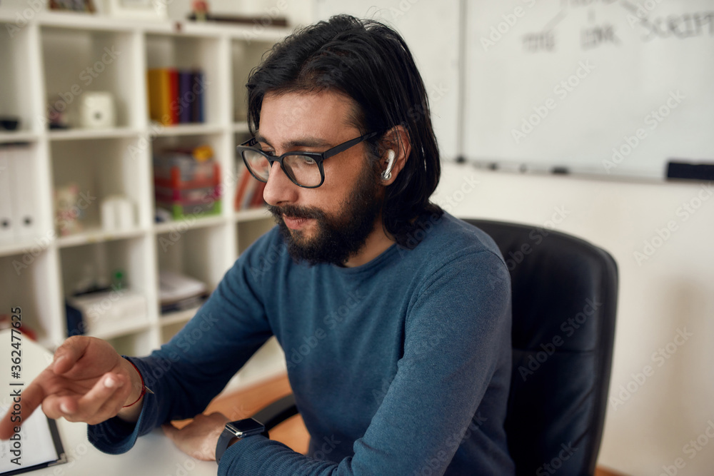 Close up of young bearded man teacher sitting at his workplace, looking at computer screen and giving online class through webcam at home Close up of young bearded man teacher sitting at his workplace, looking at computer screen and giving online class through webcam at home