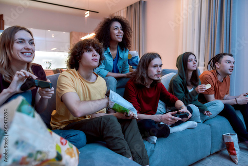 Young gamers. Young multicultural people playing video games, eating snacks and relaxing together on the couch at home. Friends spending time together