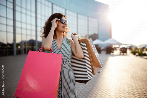 Young beautiful happy woman walks out of shopping mall with purchases. Shopaholic girl glad to buy clothes and stands outdoor with bags and credit card. Shop, sales, discount, spend money concept.