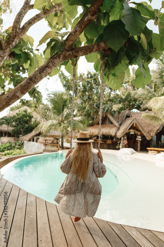 Summer vacation. Young woman wearing dress and straw hat relaxing and enjoying swing outdoors near the swimming pool. Travel and Summer Concept photo