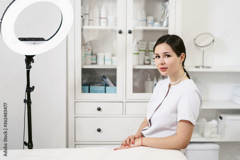 Young woman is a friendly doctor, beautician at her office in a beauty salon or cosmetology clinic. Cupboard with drugs in the background. Pure clean perfect skin and body concept. Eternal youth