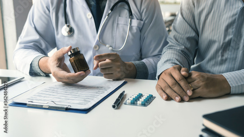 Fototapeta Naklejka Na Ścianę i Meble -  Confident doctor man holding a pill bottle and writing while talking with senior patient and reviewing his medication at office room.