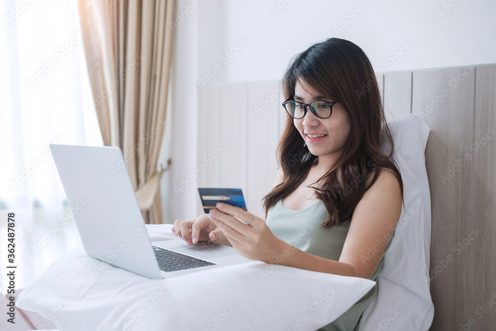 Happy woman holding credit card and using computer laptop for online shopping while making orders on bed in morning at home. Business, technology, ecommerce, digital banking and online payment concept Happy woman holding credit card and using computer laptop for online shopping while making orders on bed in morning at home. Business, technology, ecommerce, digital banking and online payment concept
