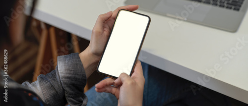 Female freelancer using mock-up smartphone while sitting at white table with laptop