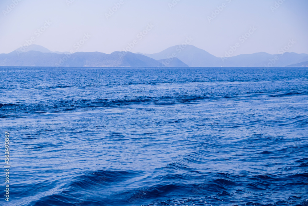 Peaceful seascape. Beautiful blue sky over calm sea with sunlight reflection. Sea waves. Mountains on horizon.