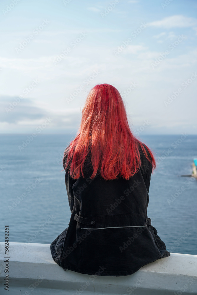 A girl with pink hair sits with her back against the sea.