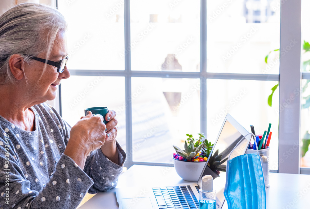 Senior woman working from home with laptop computer drinking a coffee - mask and disinfectant to protect from the coronavirus - the new normal Senior woman working from home with laptop computer drinking a coffee - mask and disinfectant to protect from the coronavirus - the new normal