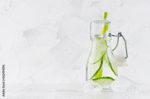 Summer homemade drink - infused green cucumber water with slices, soda, straw, ice cubes in yoke bottle in soft light white interior, copy space.