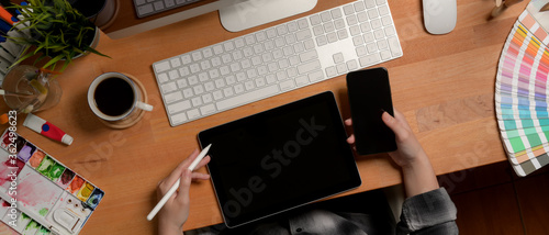 Female designer working with digital devices on office desk with painting tools and supplies