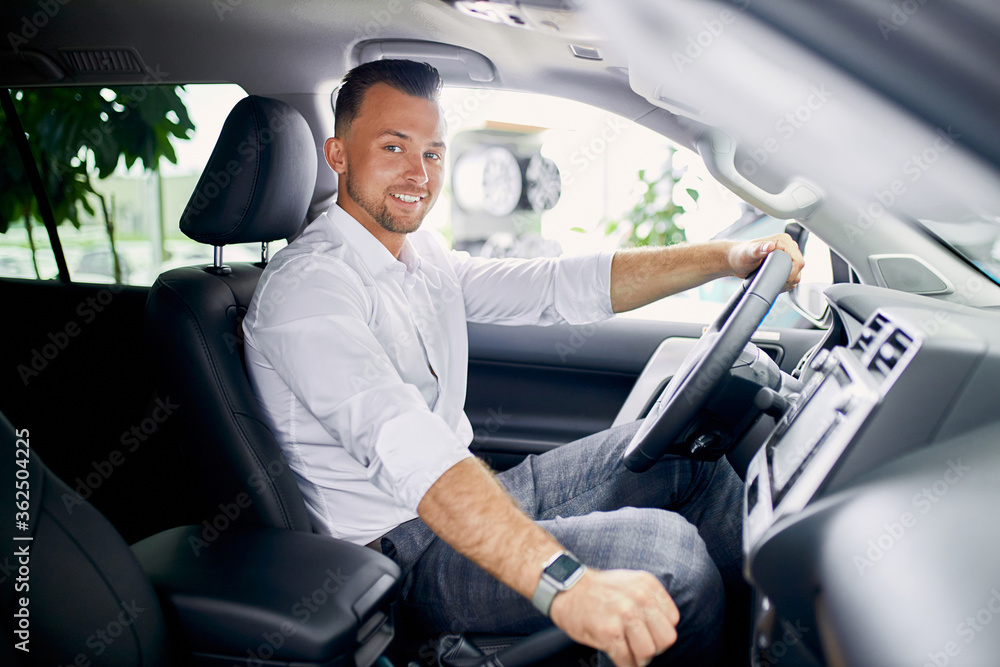 young handsome caucasian guy sits behind the wheel of new automobile, try it in driving, man is excited, going to buy car young handsome caucasian guy sits behind the wheel of new automobile, try it in driving, man is excited, going to buy car