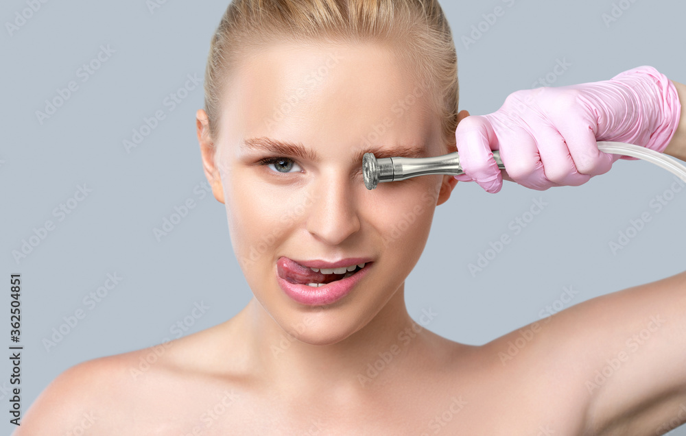 Fototapeta premium Beautiful young blonde woman with nude makeup,clean fresh skin naked shoulders holds a nozzle for microdermabrasion in her hands.She will do cosmetic procedure microdermabrasion.Aesthetic cosmetology.