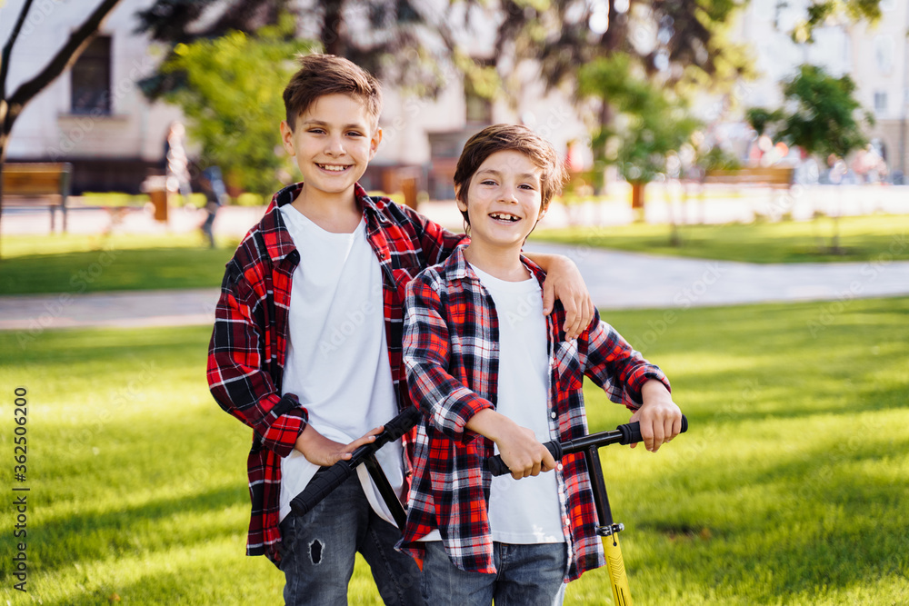 Φωτογραφία Two attractive european boys brothers, wearing red and white checkered flannel shirts, standing on scooters in the park