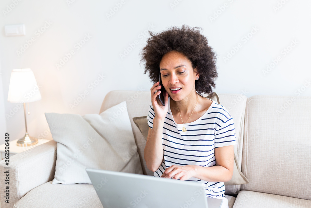 Happy woman sitting on sofa with laptop and talking on phone at home. Young successful businesswoman working from home while talking at phone. College student studying on laptop and using phone. Happy woman sitting on sofa with laptop and talking on phone at home. Young successful businesswoman working from home while talking at phone. College student studying on laptop and using phone.