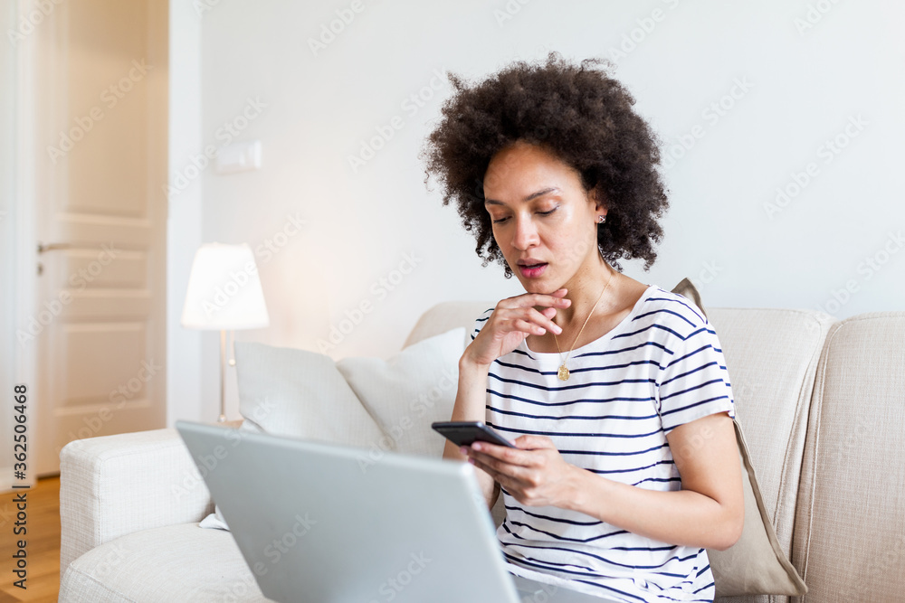 Young black woman using phone and laptop at home texting. Beautiful woman studying at home reading an sms or text message on her mobile phone with a smile as she lies on the sofa with a laptop Young black woman using phone and laptop at home texting. Beautiful woman studying at home reading an sms or text message on her mobile phone with a smile as she lies on the sofa with a laptop