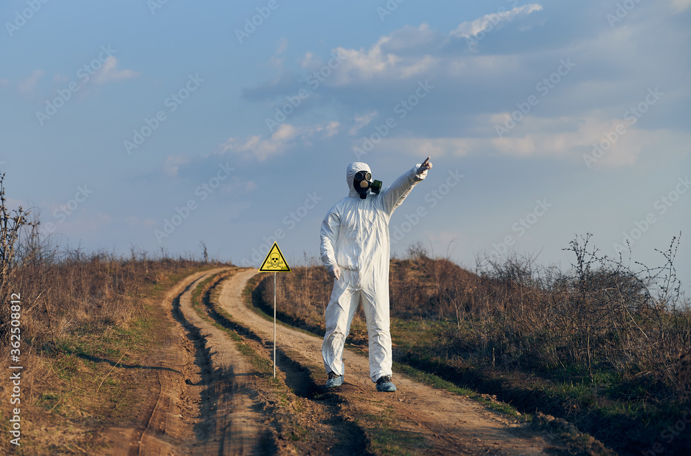 Obraz premium Environmentalist standing on the road near yellow triangle with skull and crossbones warning sign and pointing finger at sky. Research scientist wearing protective suit, gas mask and shoe covers.