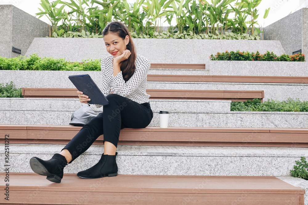 Smiling pretty young business lady sitting on wooden bench with tablet computer in hands and smiling at camera Smiling pretty young business lady sitting on wooden bench with tablet computer in hands and smiling at camera