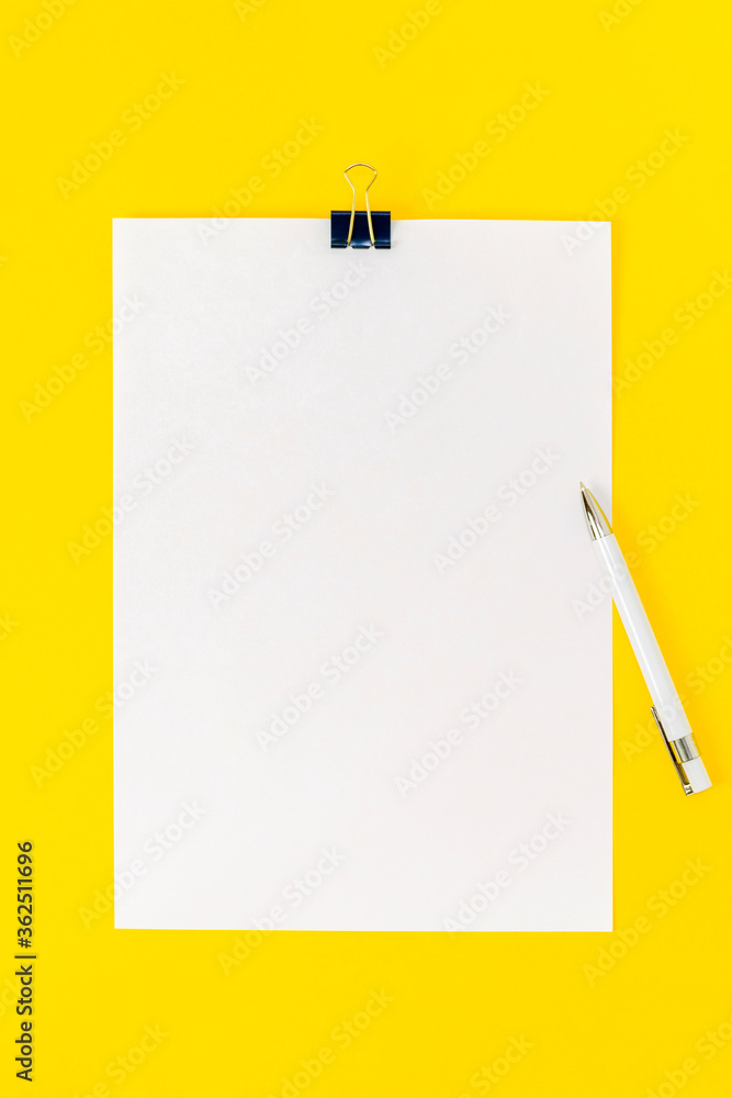 An empty sheet of paper is fastened with a stationery clip and a white pen on a yellow background. Mock up, blank for board announcement, information, statement, school. Top view, flat lay. An empty sheet of paper is fastened with a stationery clip and a white pen on a yellow background. Mock up, blank for board announcement, information, statement, school. Top view, flat lay.