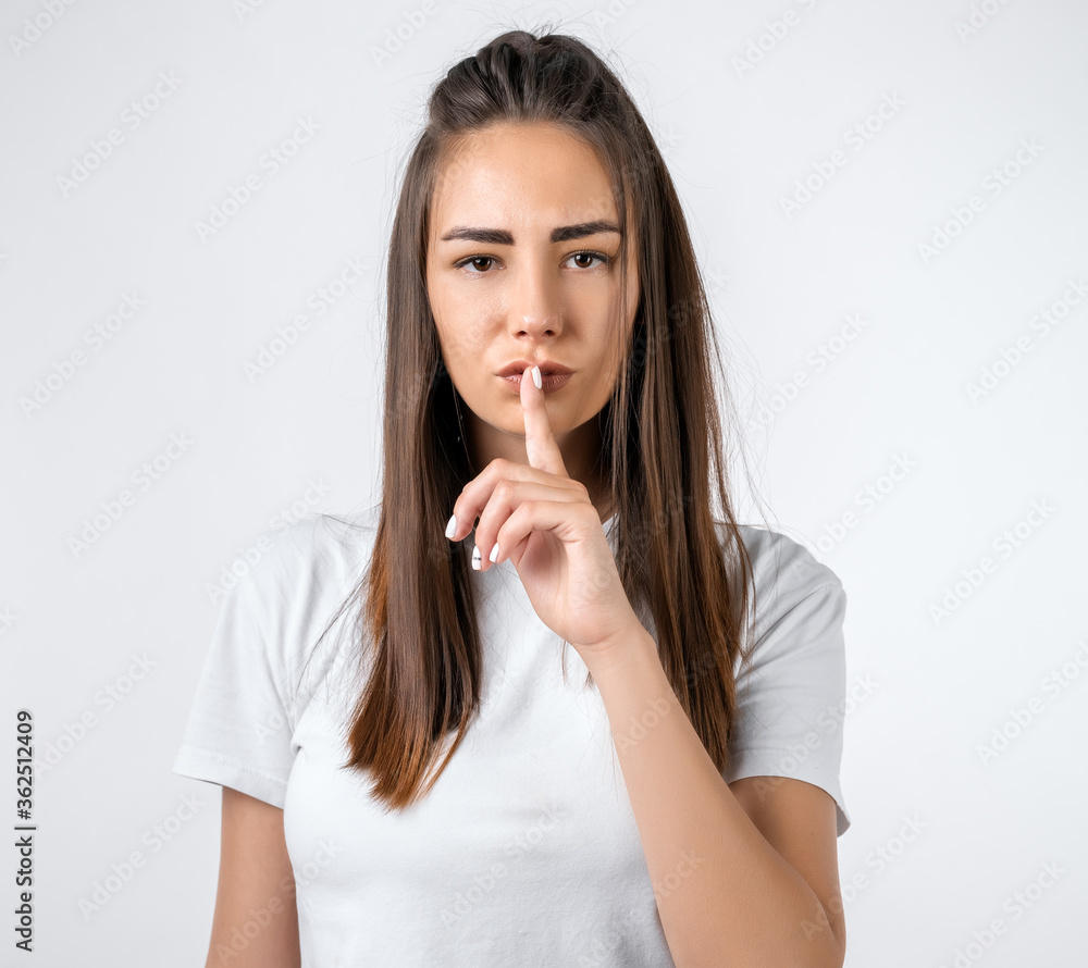 Emotional European young woman with long chestnut hair dressed in casual clothes, keeping index finger at her lips, saying Shh, Keep silent, It's secret. Emotional European young woman with long chestnut hair dressed in casual clothes, keeping index finger at her lips, saying Shh, Keep silent, It's secret.