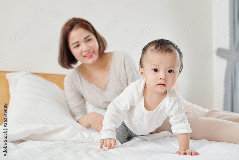 Happy young mother looking at her adorable little baby crawling on bed Happy young mother looking at her adorable little baby crawling on bed