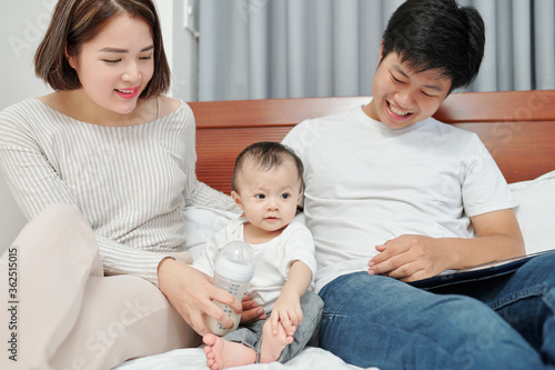 Cheerful young Asian parents sitting in bed, playing with little baby and feeding her with formula