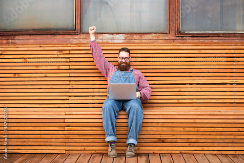 Winner. Portrait of cheerful brutal bearded hipster man in blue overalls, checked shirt working on laptop sitting on bench rising hand up celebrating attainment outdoors. Distance job, remote work. 