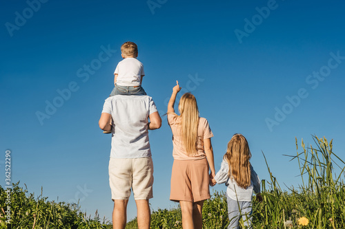Happy family of mother, father and two children have a rest in field. Mother shows a finger to the sky and they looks up. Back view.