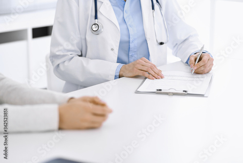 Unknown woman-doctor and female patient sitting and talking at medical examination in clinic, close-up. Therapist wearing blue blouse is filling up medication history record. Medicine concept