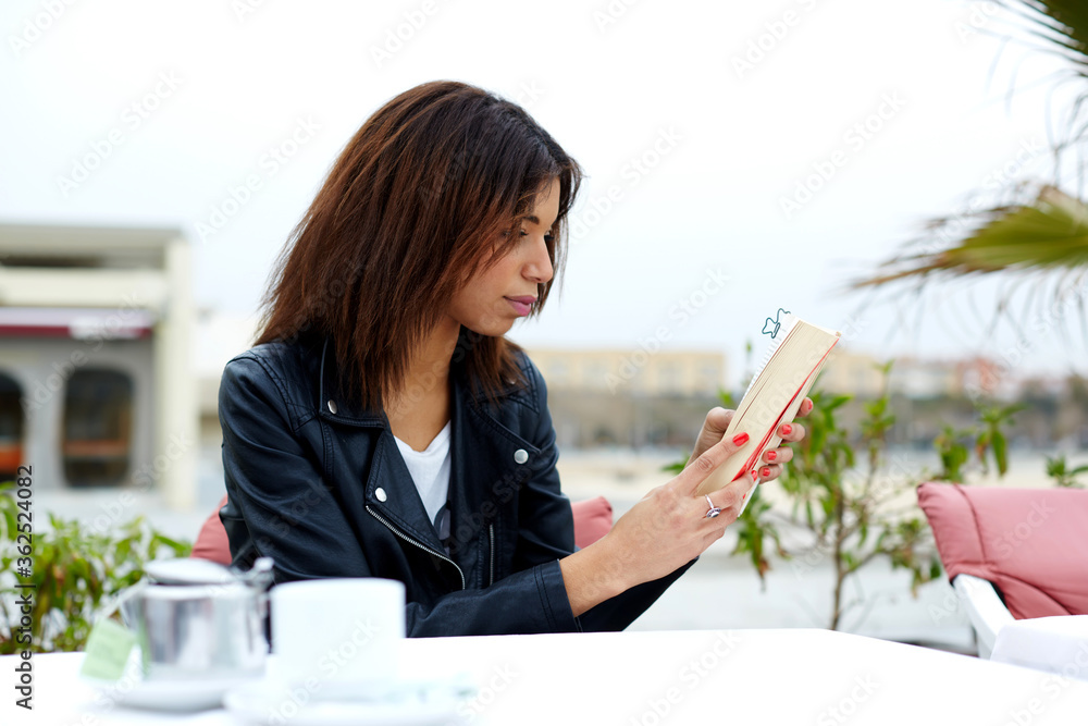 Portrait of young lovely female enjoying a good book while sitting at the table in coffee shop terrace, charming afro american woman reading novel or book during her recreation time at weekend Portrait of young lovely female enjoying a good book while sitting at the table in coffee shop terrace, charming afro american woman reading novel or book during her recreation time at weekend