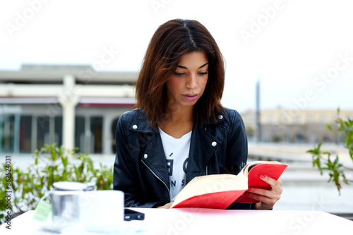 Portrait of young lovely female enjoying a good book while sitting at the table in coffee shop terrace, charming afro american woman reading novel or book during her recreation time at weekend photo