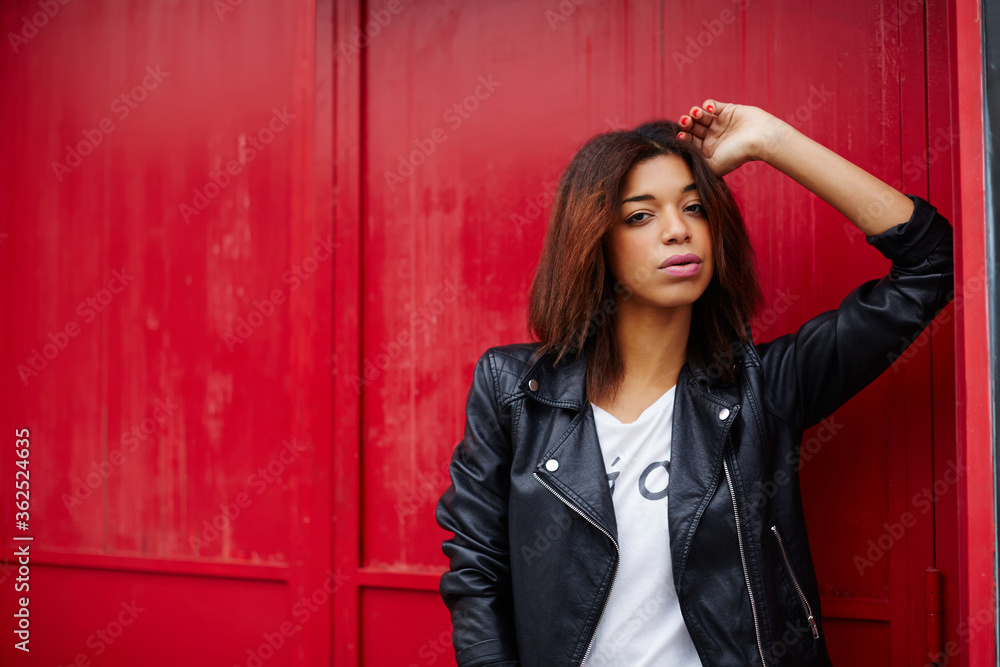 Fototapeta premium Portrait of fashionable afro american woman looking to you, attractive black female posing on red wall background in the city, cute hipster clothing