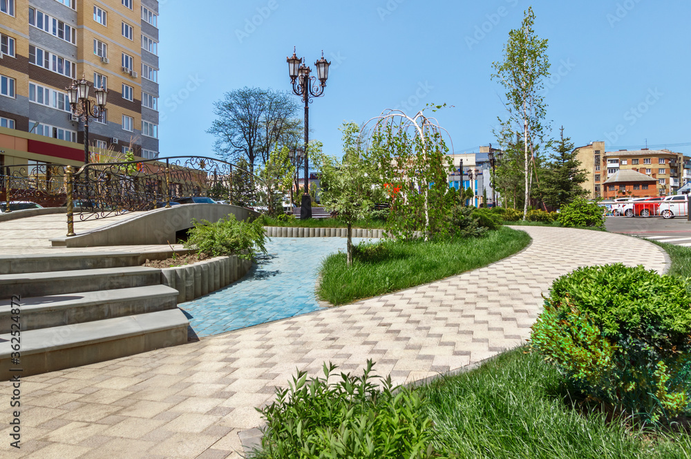 Fototapeta premium The small park near a residential buildings with the paved stone walkways, the lawn and the decorative bridge