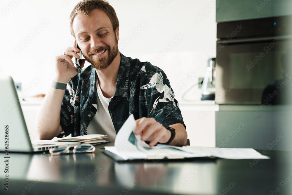 Photo of smiling man talking on cellphone while working with laptop Photo of smiling man talking on cellphone while working with laptop