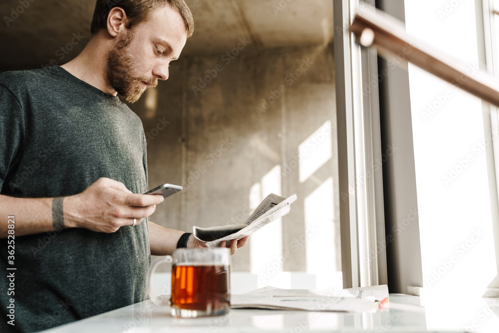Photo of serious redhead man reading newspaper while using mobile phone Photo of serious redhead man reading newspaper while using mobile phone