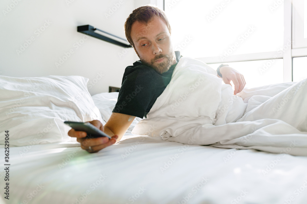 Photo of young bearded man using mobile phone while lying after sleep Photo of young bearded man using mobile phone while lying after sleep