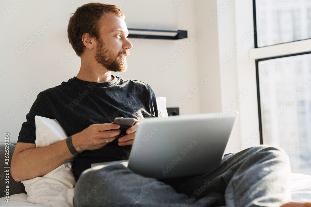 Photo of focused man using cellphone and laptop while sitting on bed Photo of focused man using cellphone and laptop while sitting on bed
