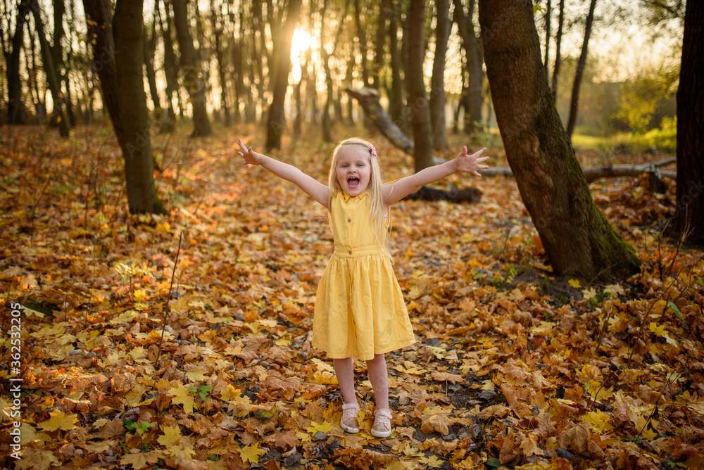 Little blonde girl in a yellow dress in the autumn park. The child holds his hands in different directions and enjoys a warm autumn.