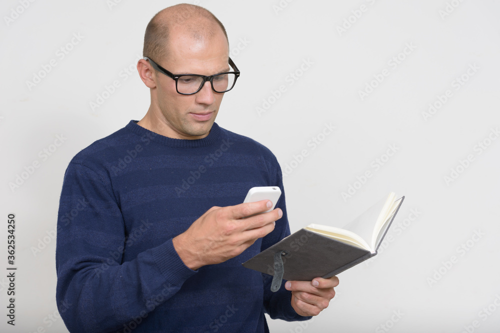 Portrait of young bald nerd man using phone while holding book Portrait of young bald nerd man using phone while holding book