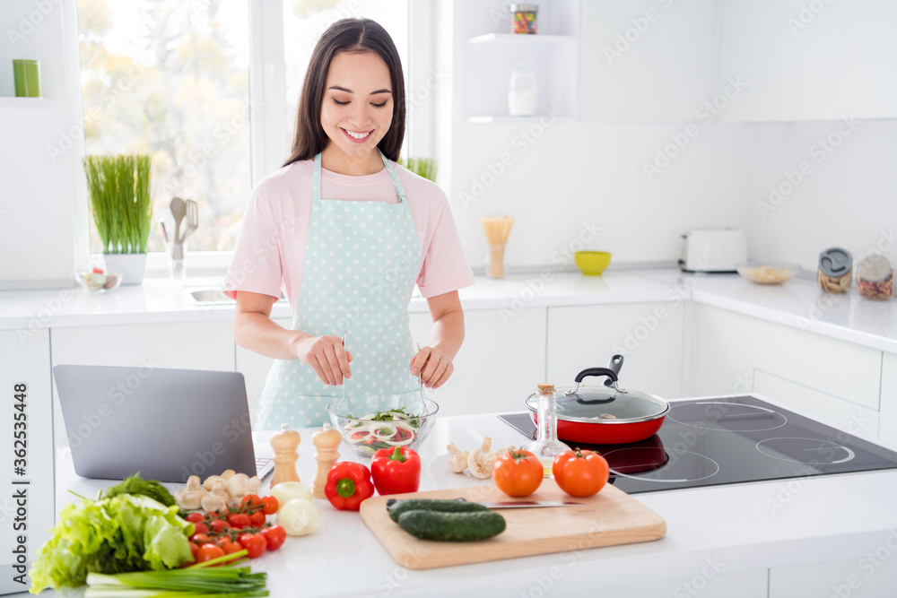 Photo of asian ethnicity housewife mixing fresh vegetables salad ingredients cooking tasty vegan meal using online blogger recipe notebook on table stand kitchen indoors