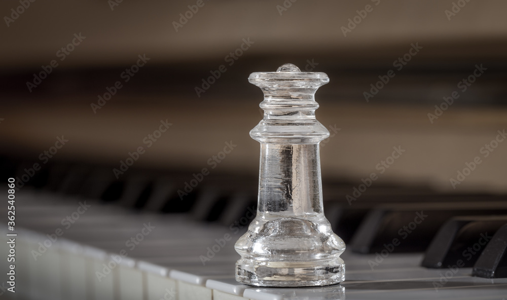 Clear glass chess figure - King/Queen - standing on the keys of an old wooden piano. Closeup shot with big commercial space and wide range of possible use. Focus on bishop, background out of focus. Clear glass chess figure - King/Queen - standing on the keys of an old wooden piano. Closeup shot with big commercial space and wide range of possible use. Focus on bishop, background out of focus.
