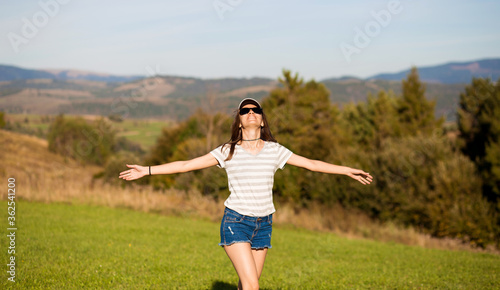 Beautiful carefree woman in sunglasses, cap and jeans shorts walk in summer field with mountains