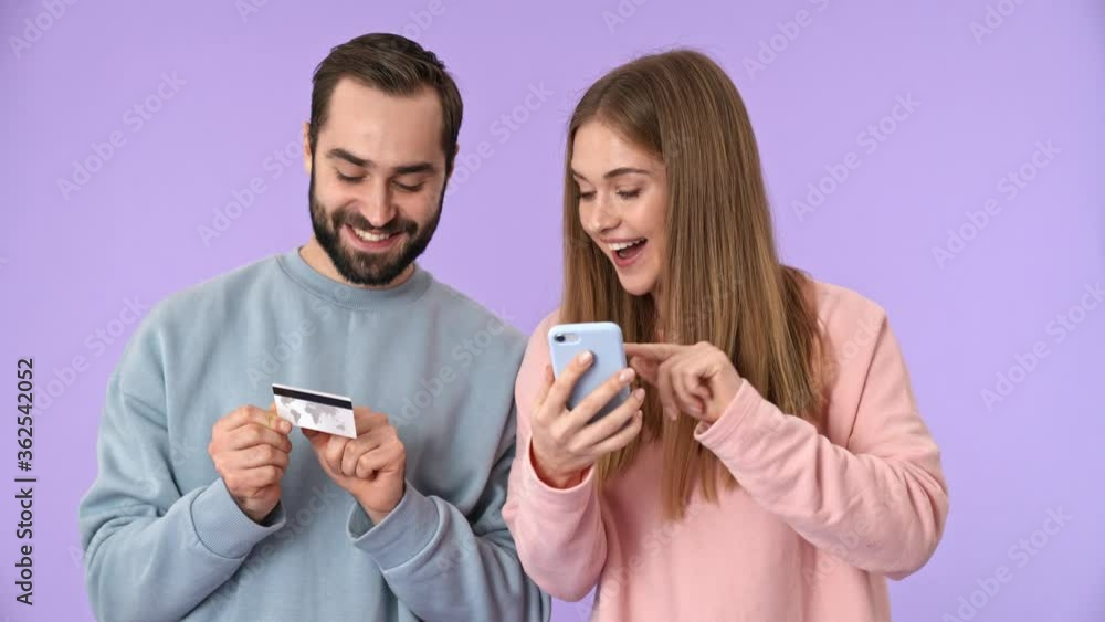 Cheerful young couple making a purchases on smartphone with credit card and rejoicing over purple background Cheerful young couple making a purchases on smartphone with credit card and rejoicing over purple background