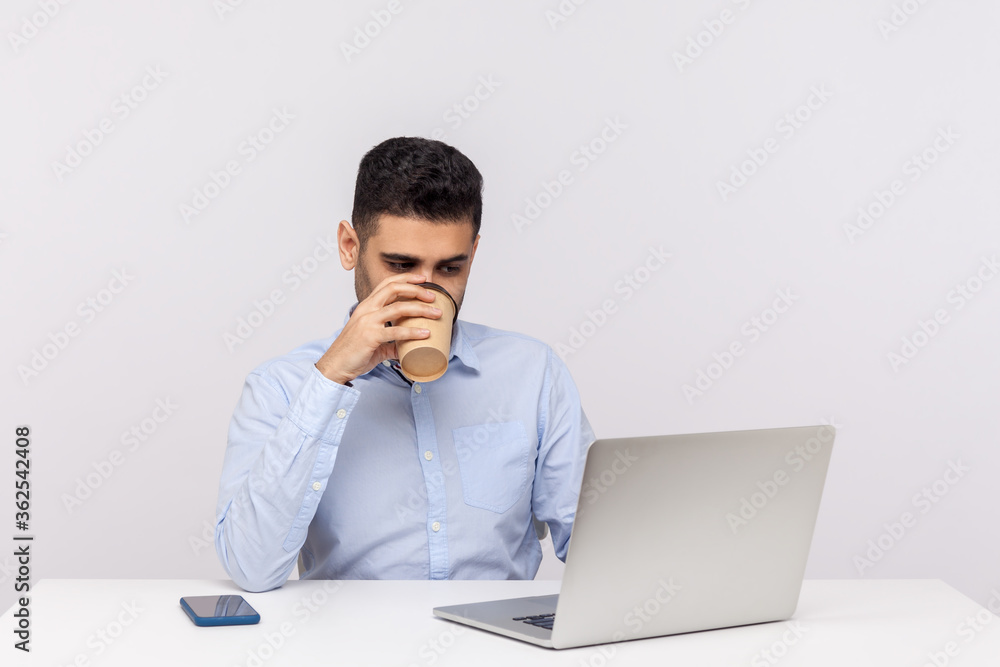 Busy elegant young man employee sitting in office workplace, working on laptop to get all his business done early in morning with cup of coffee. indoor studio shot isolated on white background Busy elegant young man employee sitting in office workplace, working on laptop to get all his business done early in morning with cup of coffee. indoor studio shot isolated on white background