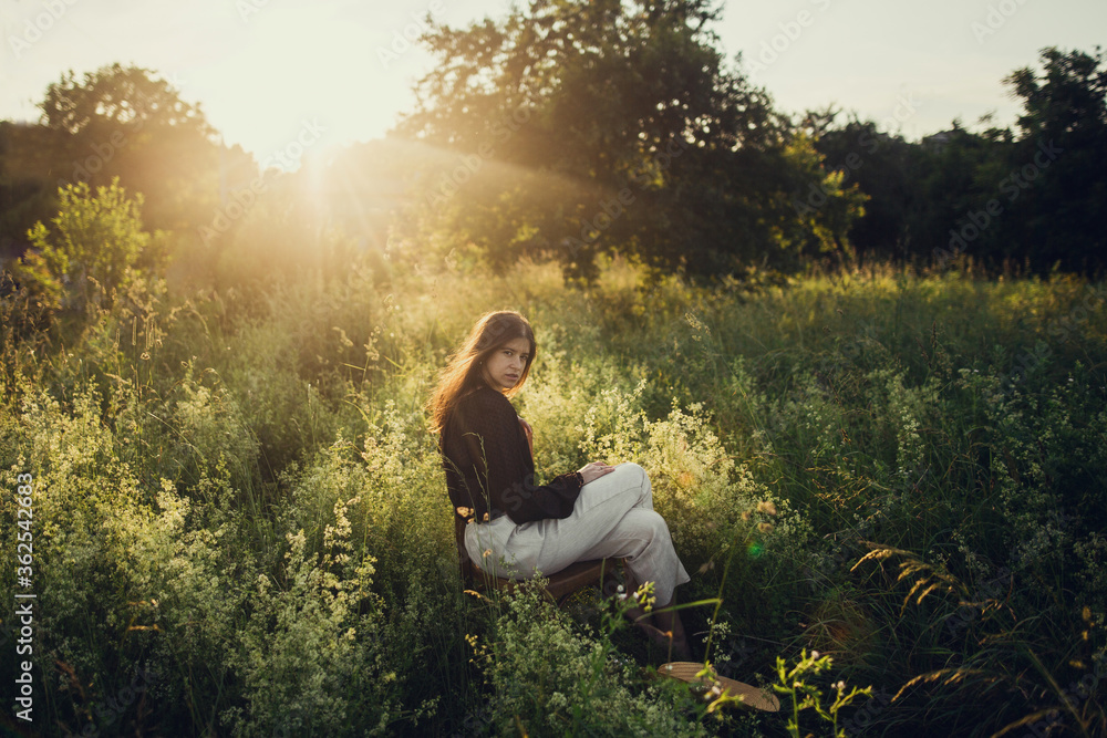 Fashionable woman relaxing in summer countryside. Stylish elegant girl sitting on rustic chair in sunset light in summer meadow.Atmospheric tranquil moment. Creative image. Fashionable woman relaxing in summer countryside. Stylish elegant girl sitting on rustic chair in sunset light in summer meadow.Atmospheric tranquil moment. Creative image.