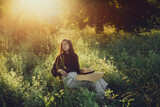 Stylish elegant girl sitting on rustic chair in sunset light in summer meadow. Fashionable woman relaxing in summer countryside. Creative image. Atmospheric tranquil moment