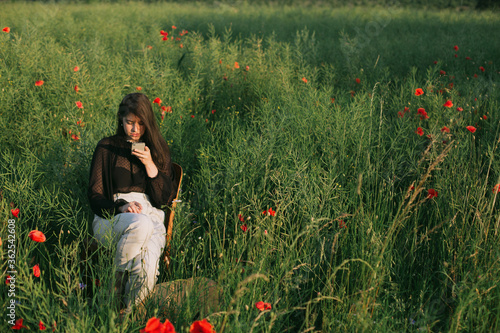 Stylish elegant girl chatting online, holding phone and sitting in summer meadow in warm sunset light. Fashionable young woman using smartphone making photos © sonyachny