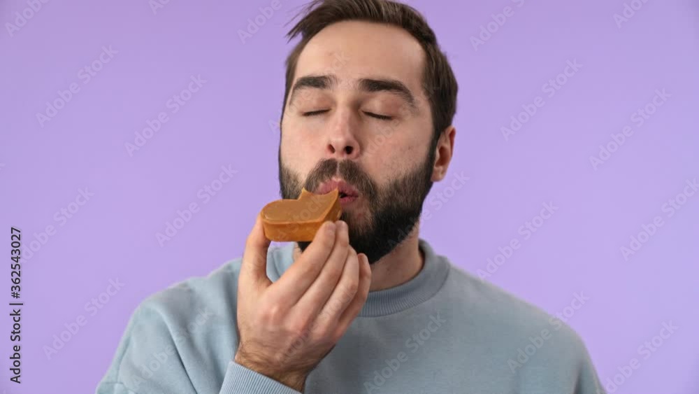Smiling bearded man showing cake at the camera then eating it and enjoying the moment over purple background Smiling bearded man showing cake at the camera then eating it and enjoying the moment over purple background